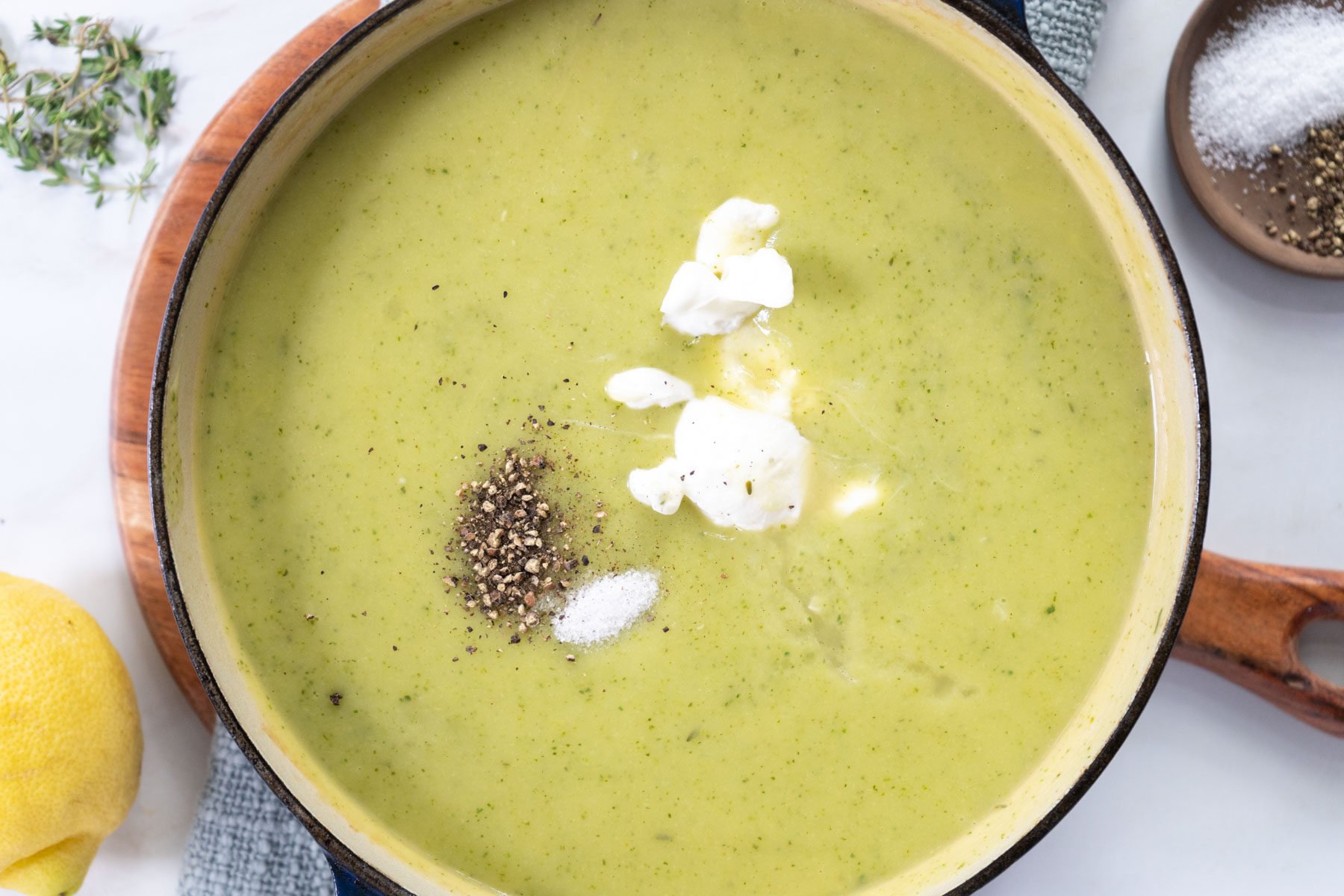 A pot of creamy zucchini soup topped with three dollops of cream, a small pile of ground black pepper, and a pinch of salt. Fresh thyme, a lemon, and a small bowl of pepper are nearby on a white surface.