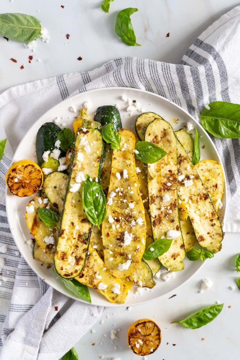 Grilled zucchini and squash on a plate with a dish towel and grilled lemons.