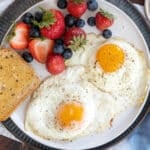 Two sunny side up eggs on a plate with toast and fruit.