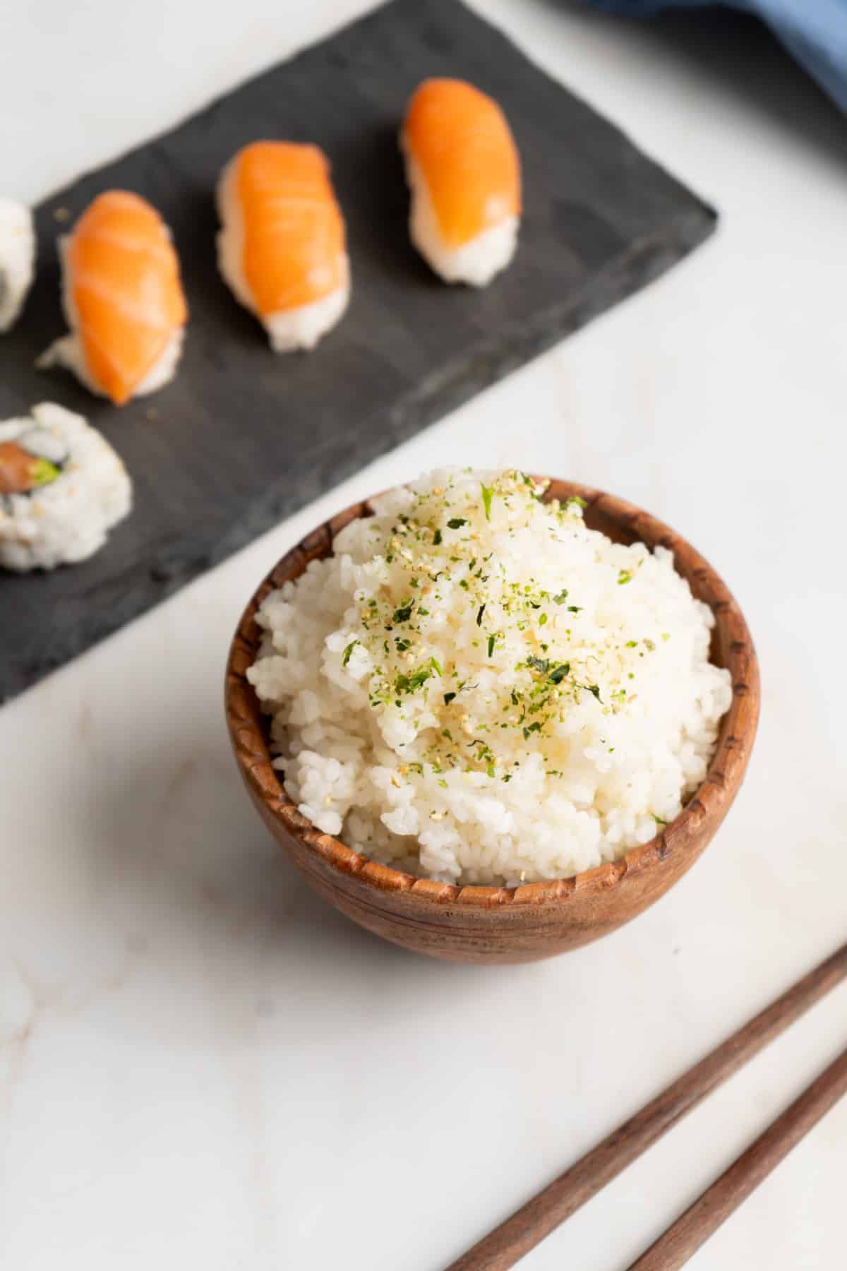Sushi rice in a wood bowl with furikake seasoning and a plate of nigiri.