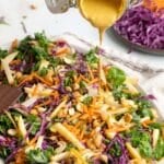A colorful kale apple slaw with shredded carrots, red cabbage, apple slices, and cashews is being drizzled with a creamy dressing.