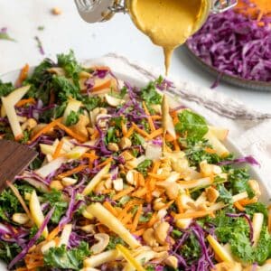 A colorful kale apple slaw with shredded carrots, red cabbage, apple slices, and cashews is being drizzled with a creamy dressing.