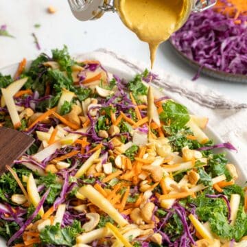 A colorful kale apple slaw with shredded carrots, red cabbage, apple slices, and cashews is being drizzled with a creamy dressing.