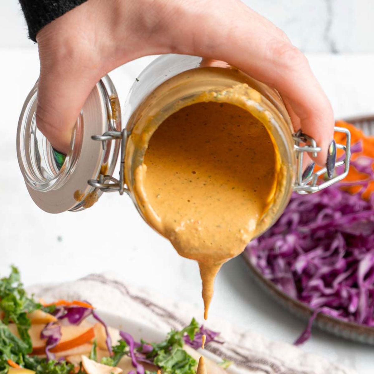 A hand pours creamy Cashew Dressing from a glass jar over a plate of salad with kale, carrots, and shredded purple cabbage.