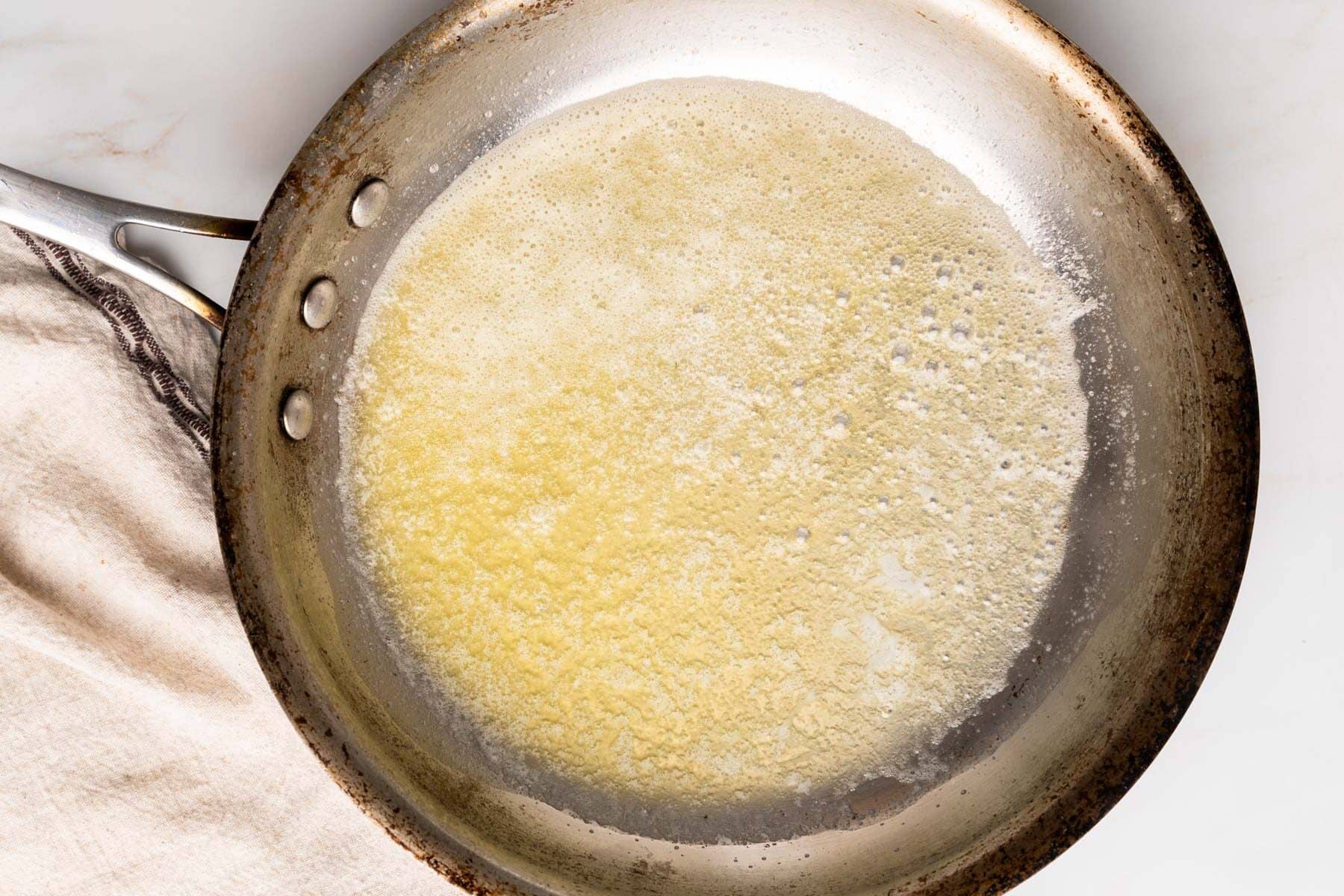 A frying pan on a stove with melted butter covering the bottom, ready for creating a gluten-free roux. A cloth is partially visible on the side.