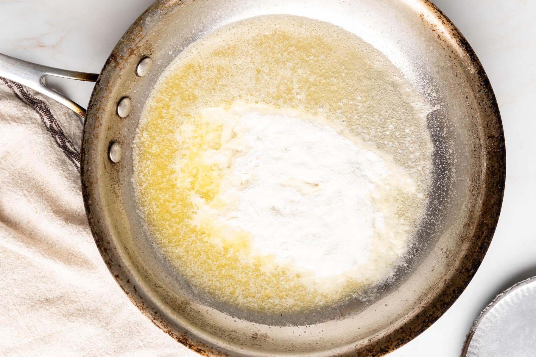 A gluten-free roux of butter and flour mixture gently melts in a stainless steel pan on the stovetop, with a cloth nearby.