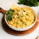 A bowl of creamy Cajun alfredo pasta garnished with grated cheese, chopped herbs, microgreens, and a lemon wedge, placed on a wooden board with fresh parsley in the background.
