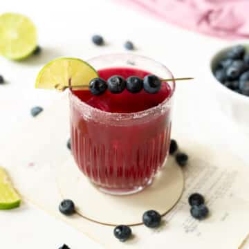 A glass of blueberry margarita garnished with a fruit skewer of blueberries with a slice of lime on the rim.⁣
