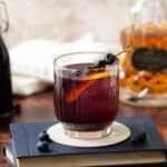 A glass filled with blueberry syrup, ice, and a lemon slice, garnished with fresh blueberries on small fruit picks, resting atop a book on a countertop, with a bottle of whiskey in the background.