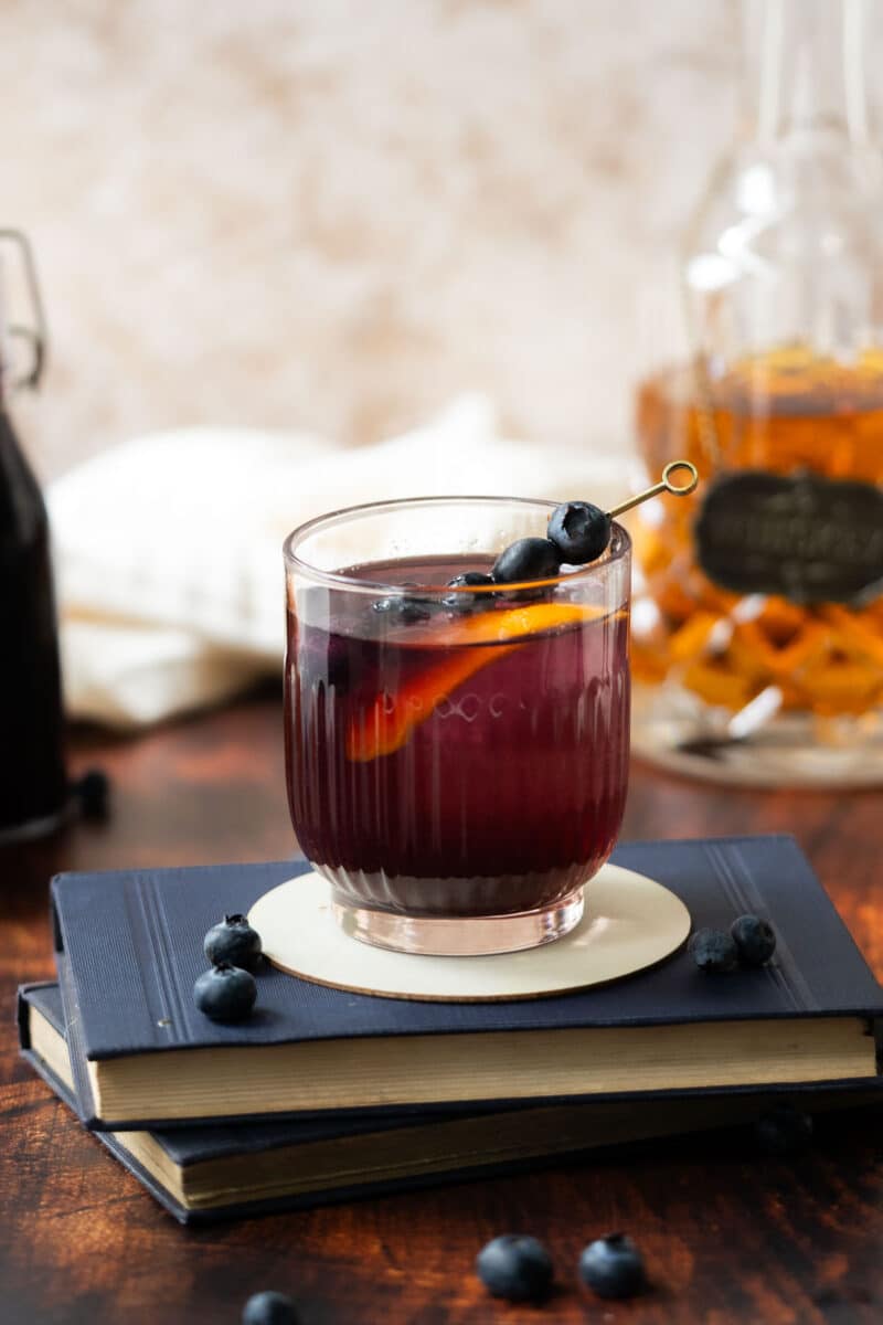 A glass filled with blueberry syrup, ice, and a lemon slice, garnished with fresh blueberries on small fruit picks, resting atop a book on a countertop, with a bottle of whiskey in the background.