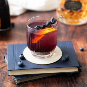 A glass filled with blueberry syrup, ice, and a lemon slice, garnished with fresh blueberries on small fruit picks, resting atop a book on a countertop, with a bottle of whiskey in the background.