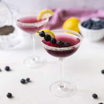 A blueberry martini glass with blueberries on fruit skewers, half dipped in the drink, and a lemon slice on the rim, with an additional martini glass in the background, adorned similarly.⁣