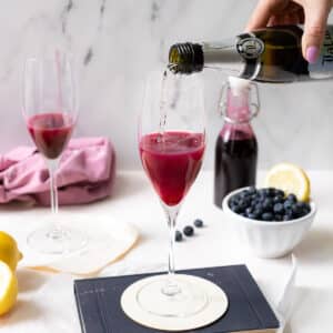 A champagne glass with blueberry syrup poured with prosecco wine into it, with a bottle and a bowl of blueberries with a slice of lemon on the side.⁣⁣⁣⁣