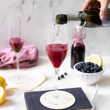 A champagne glass with blueberry syrup poured with prosecco wine into it, with a bottle and a bowl of blueberries with a slice of lemon on the side.⁣⁣⁣⁣