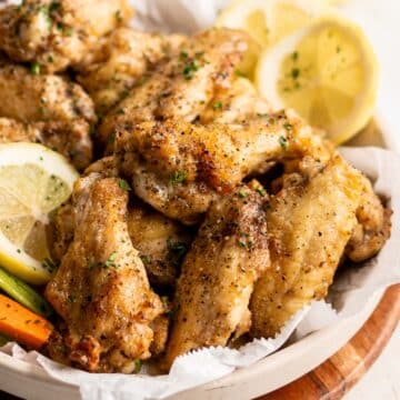 Lemon pepper chicken wings, adorned with minced parsley, lemon slices, carrot batons, and fresh parsley sprigs, on a rustic wooden platter.⁣