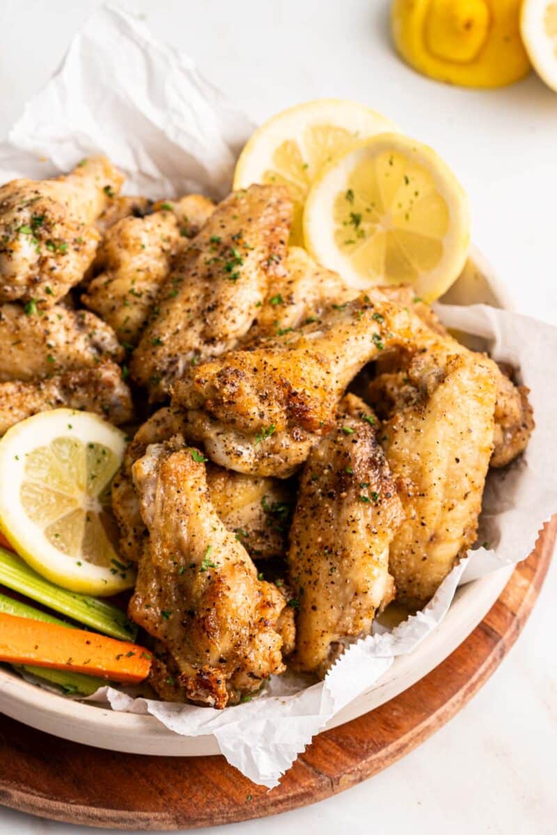 Lemon pepper chicken wings, adorned with minced parsley, lemon slices, carrot batons, and fresh parsley sprigs, on a rustic wooden platter.⁣