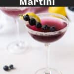 A blueberry martini with blueberries on fruit skewers partially dipped in the drink, along with a lemon slice on the rim.