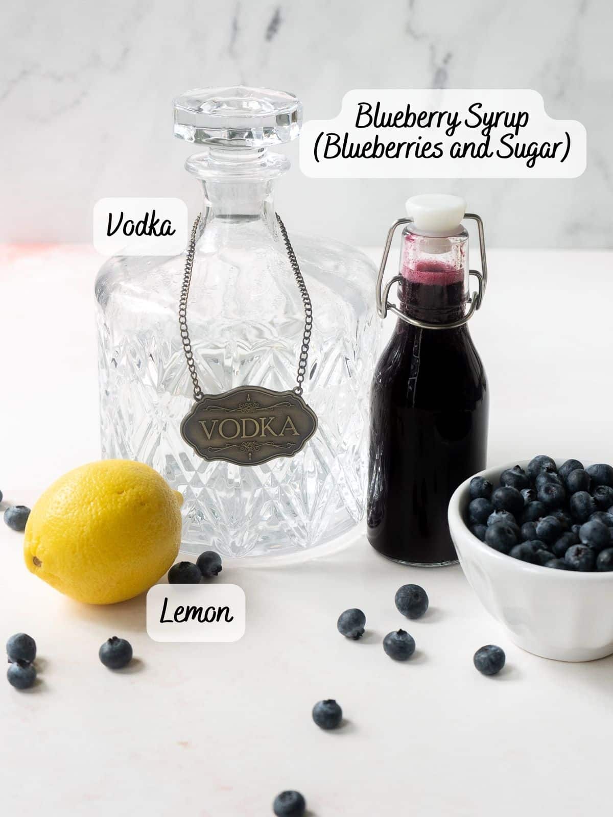 The image displays the ingredients for a blueberry martini: lemon, vodka bottle, blueberry syrup bottle, and a bowl of blueberries.