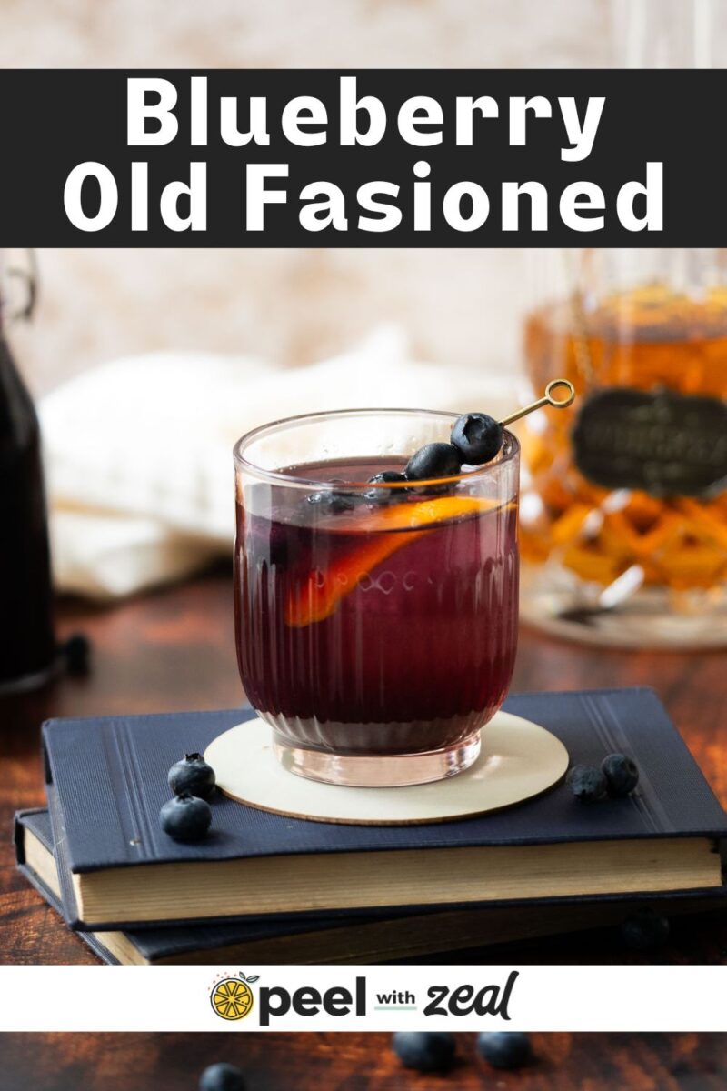 A glass filled with blueberry syrup, ice, and a lemon slice, garnished with fresh blueberries on small fruit picks, resting atop a book on a countertop, with a bottle of whiskey in the background.