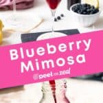 A champagne glass with blueberry syrup and prosecco being poured into it, making a Blueberry Mimosa, with some blueberries and a lemon slice on the side.