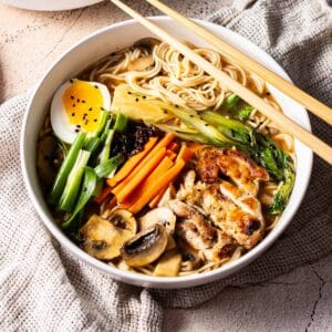 Bowl of ramen served on the bowl garnish with batons of carrots, scallions, halves of boiled egg, mushroom and chicken meat place on the table.