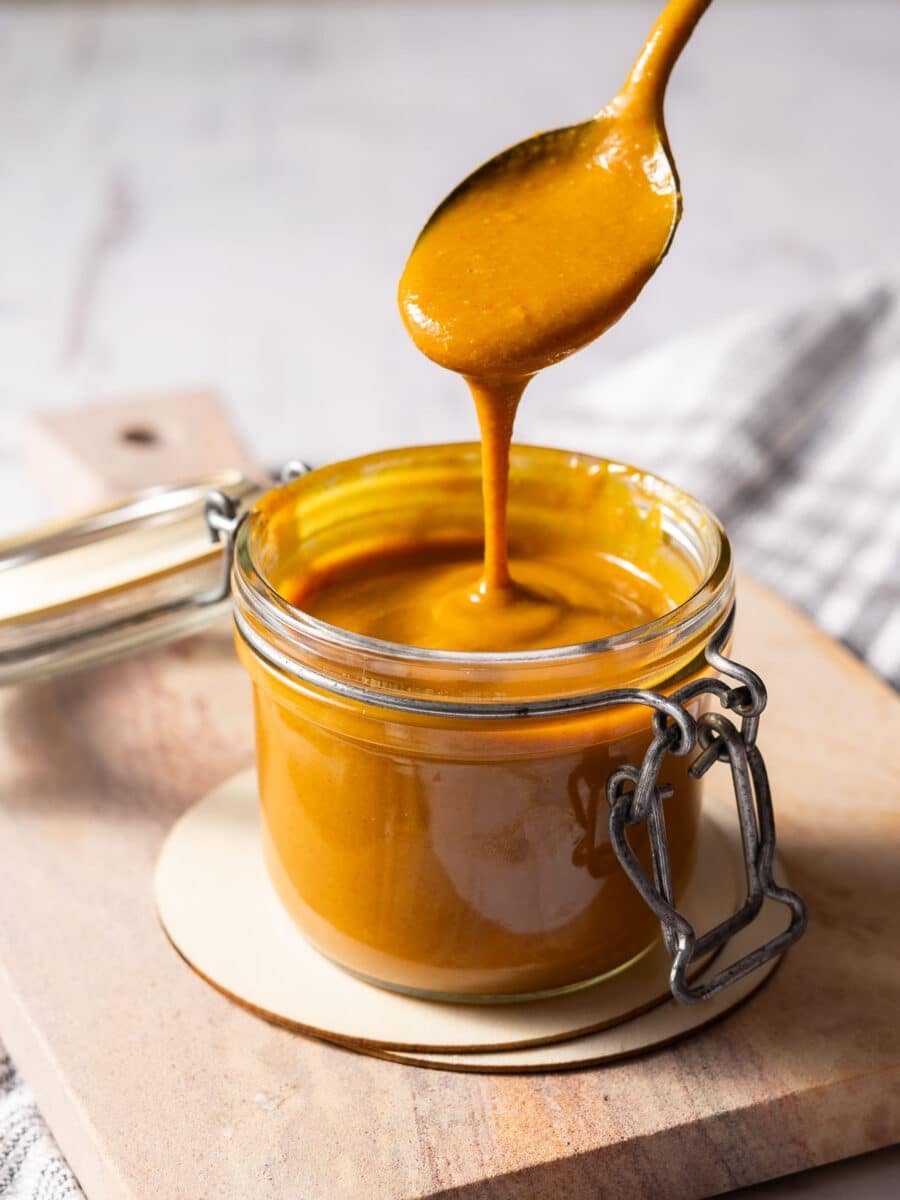 A spicy mustard BBQ sauce dripping from the spoon filling up the jar on the board atop the table.