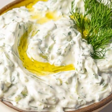 A bowl of creamy cold dip garnished with fresh dill and a drizzle of olive oil, perfect for cookouts or to chill out with friends, served on a plate.