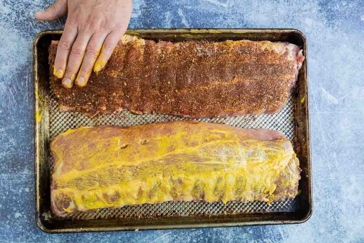 Two racks of ribs, one covered with mustard and the other with dry rub. 