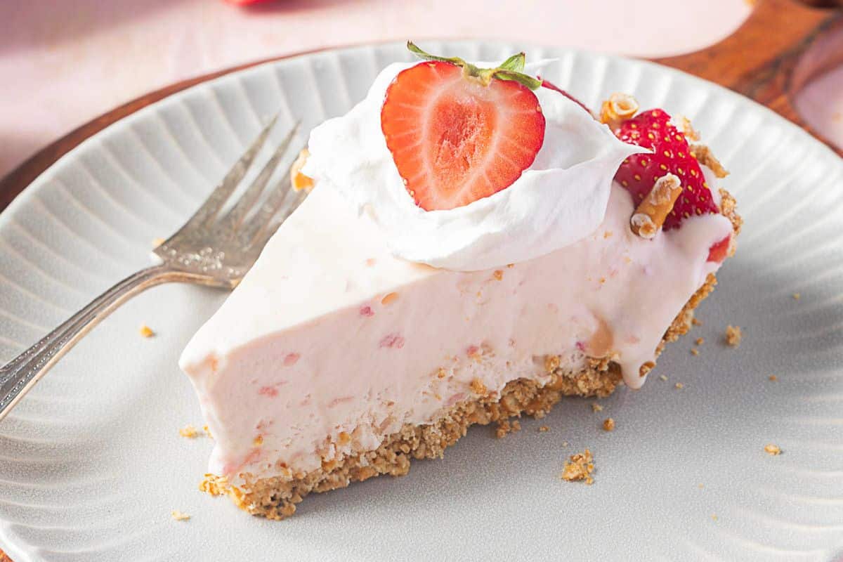 A slice of strawberry cream pie with a graham cracker crust, topped with whipped cream and a halved strawberry, served on a plate with a fork—one of the most irresistible desserts and perfect for summer recipes.