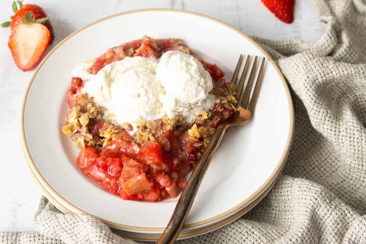 A plate with strawberry-rhubarb crisp topped with two scoops of vanilla ice cream, a fork, and a sliced strawberry on the side—one of those irresistible desserts perfect for summer recipes.