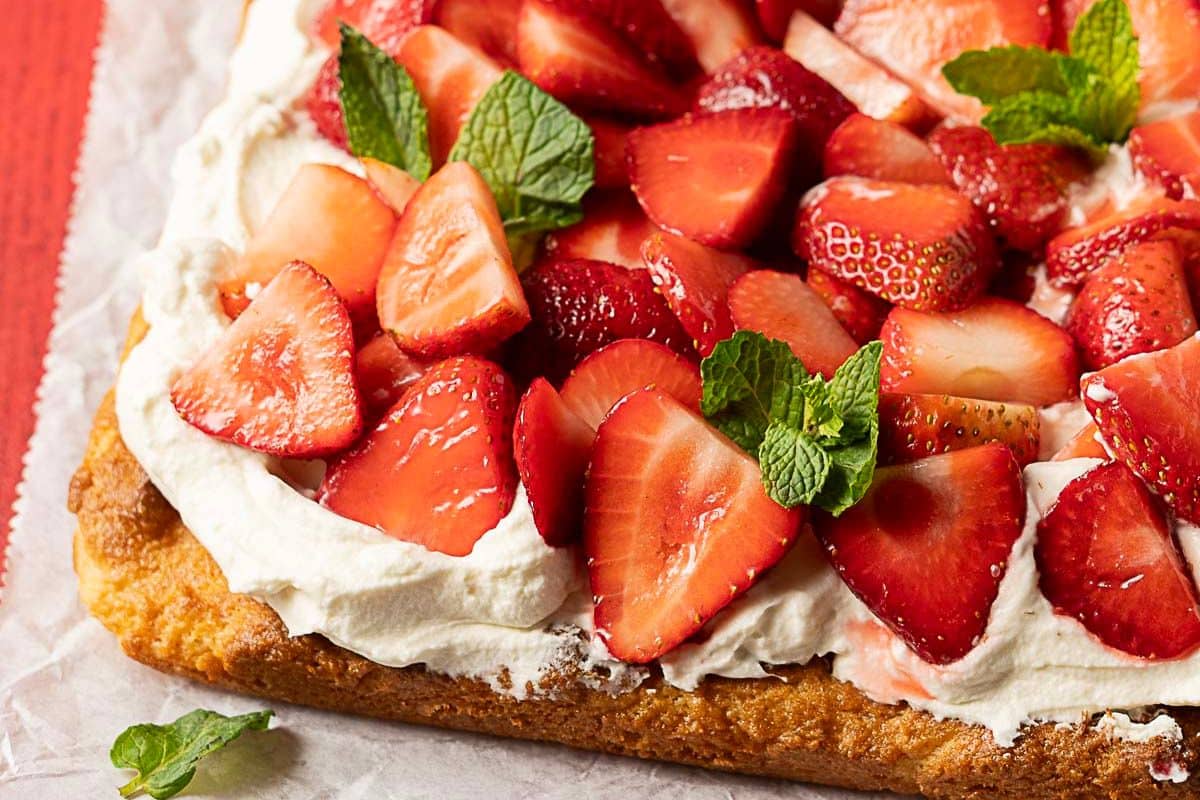 A close-up of a rectangular sponge cake topped with whipped cream, sliced fresh strawberries, and mint leaves on parchment paper—an irresistible treat perfect for summer desserts or your favorite strawberry recipes.