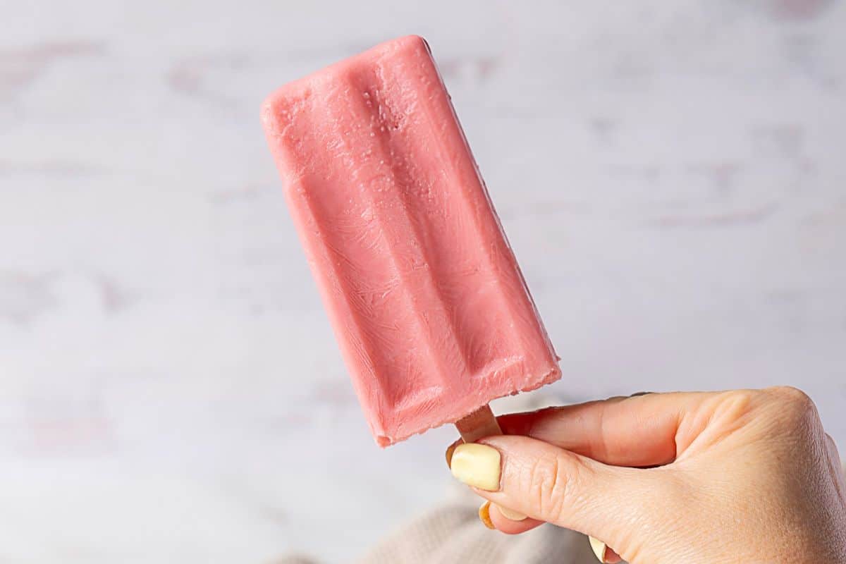 A hand holds a pink popsicle on a wooden stick against a light, blurred background—one of those irresistible desserts perfect for summer recipes.