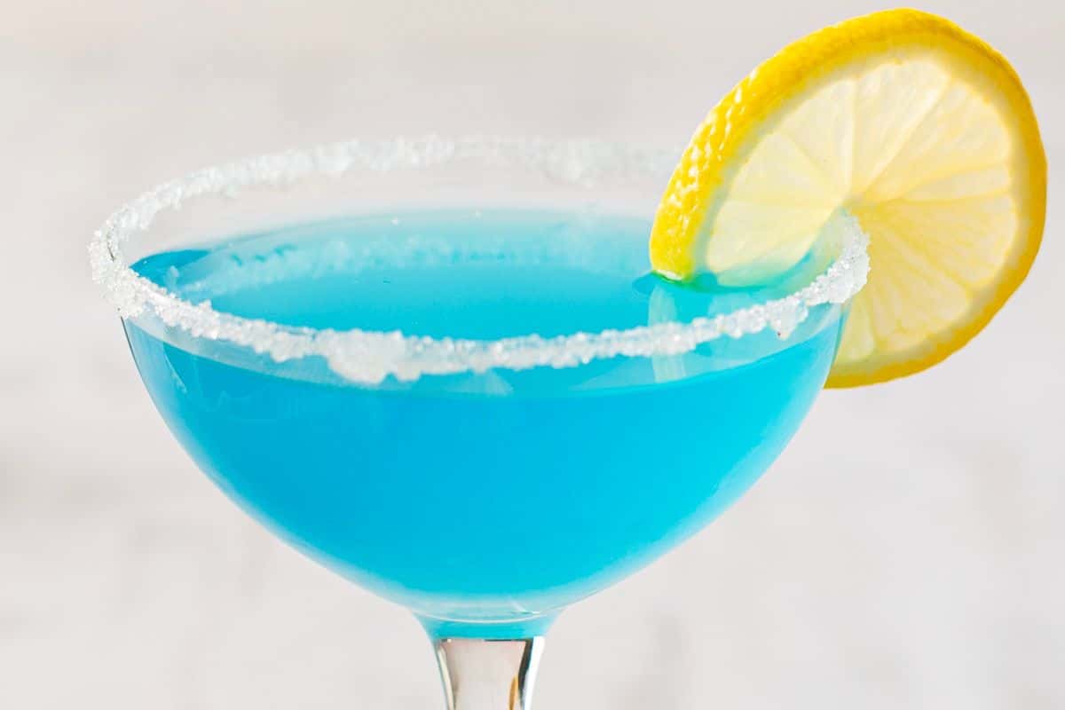 A blue cocktail in a glass with a sugared rim, garnished with a slice of lemon—perfect for summer cocktails and refreshing Memorial Day cocktails.