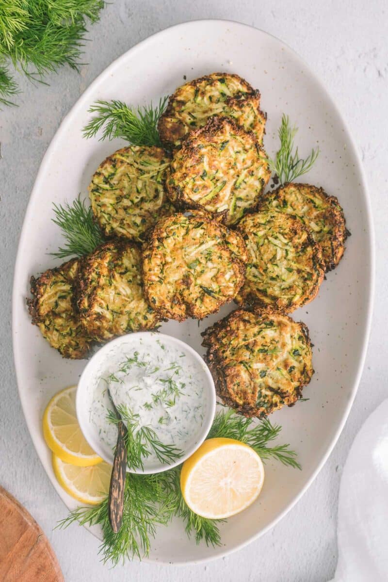 Zucchini fritters served on a white oval shape platter with dill sauce .
