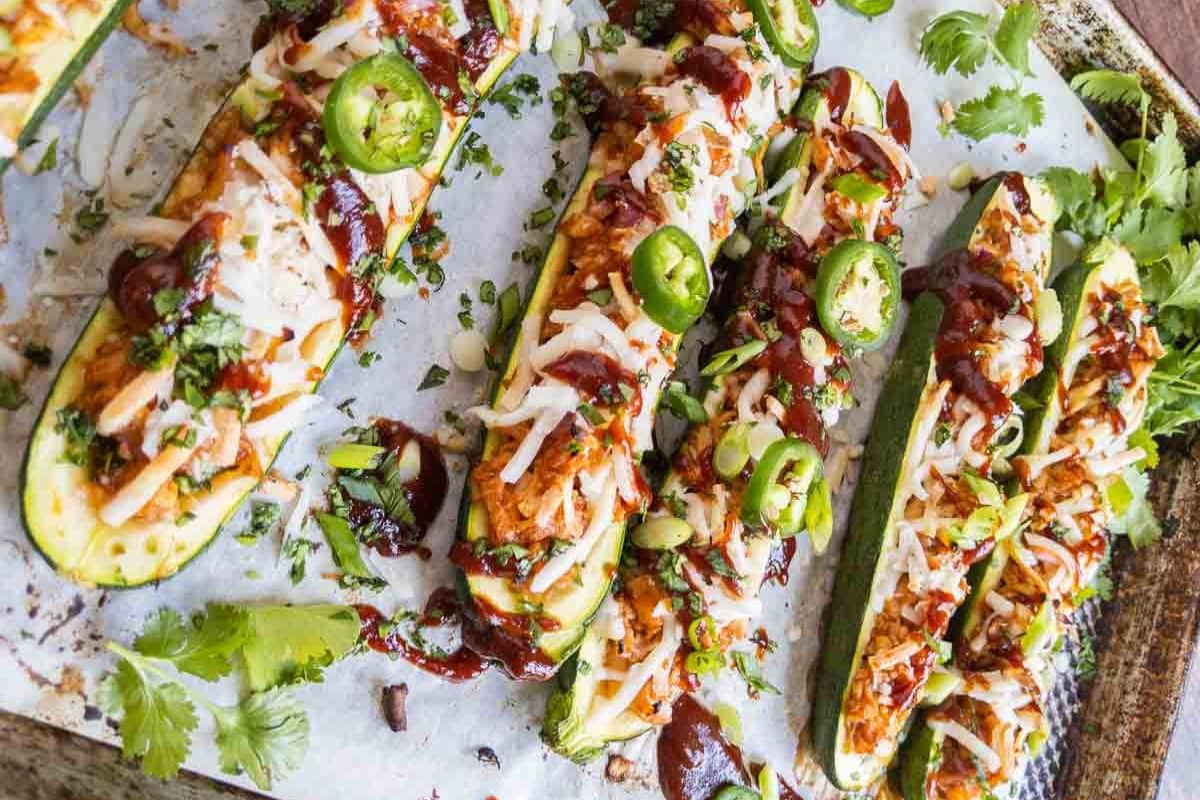 Zucchini halves topped with shredded cheese, barbecue sauce, jalapeño slices, and chopped herbs—this is one of the most creative zucchini recipes for summer, arranged on a parchment-lined baking sheet.