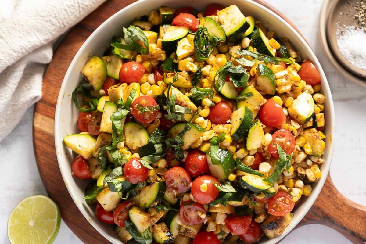A bowl of grilled corn salad with cherry tomatoes, zucchini, fresh basil, and spices on a wooden board, next to a lime wedge and a towel—perfect for anyone seeking creative zucchini recipes for their summer menu.