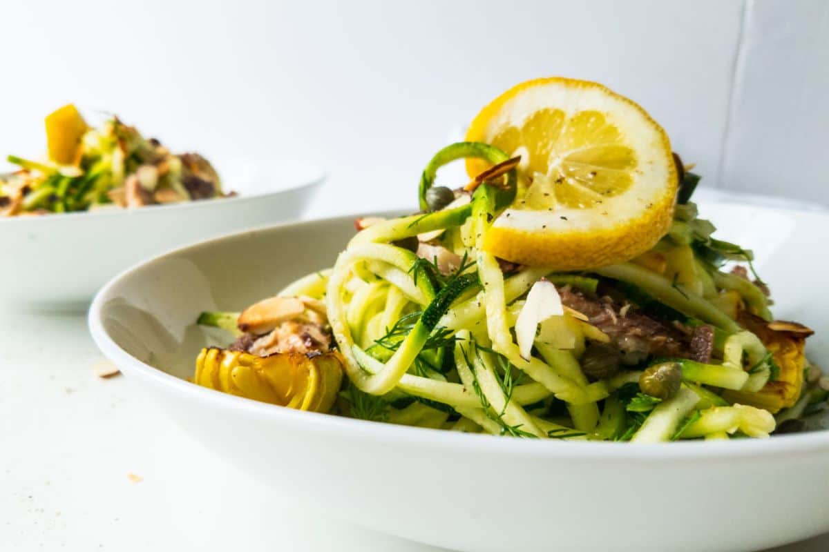 A white bowl of zucchini noodles topped with lemon slices, herbs, capers, and vegetables, with another similar bowl in the background—perfect for those seeking creative zucchini recipes on a bright white surface.