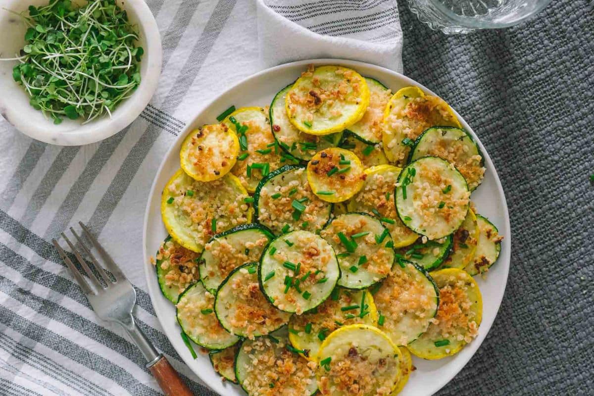 A plate of baked zucchini and yellow squash slices topped with breadcrumbs and chopped chives, placed on a striped cloth next to a bowl of sprouts and a fork—perfect for trying out creative zucchini dishes and fresh summer recipes.