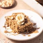 A portion of Baked Oatmeal Peanut Butter topped with slices of banana and nuts drizzled with peanut sauce served on a plate.