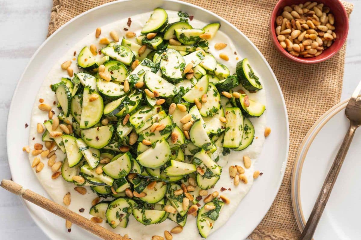 A plate of sliced zucchini mixed with herbs and toasted pine nuts sits on a creamy white spread, perfect for those seeking creative zucchini recipes, with a small bowl of pine nuts and utensils beside it.