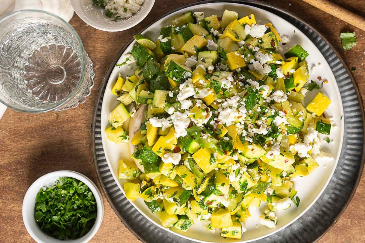 A plate of diced yellow and green squash mixed with herbs and crumbled cheese, next to a glass of water and a small bowl of chopped parsley on a wooden table—perfect for those seeking creative zucchini recipes.