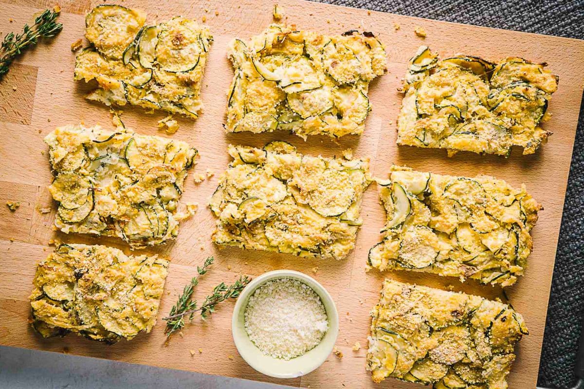 Sliced zucchini casserole on a wooden cutting board with a small bowl of grated cheese and fresh thyme nearby—perfect for those seeking creative zucchini recipes.