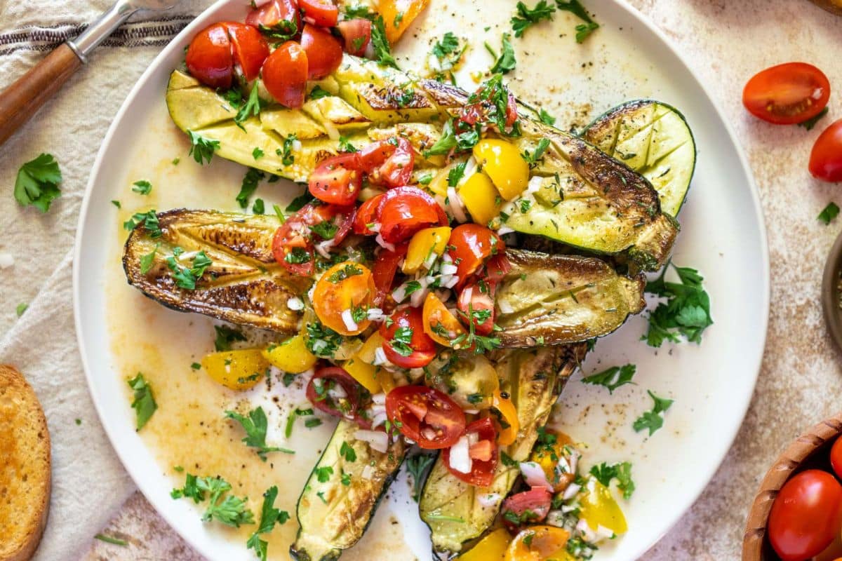 Grilled zucchini halves topped with chopped tomatoes, yellow bell peppers, onions, and fresh herbs make this a standout among creative zucchini recipes for summer. Served beautifully on a white plate.