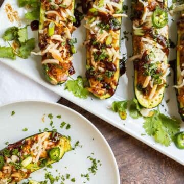 A plate and tray of zucchini boats stuffed with shredded cheese, barbecue sauce, sliced jalapeños, and chopped herbs, garnished with fresh cilantro—perfect for creative zucchini recipes this summer.