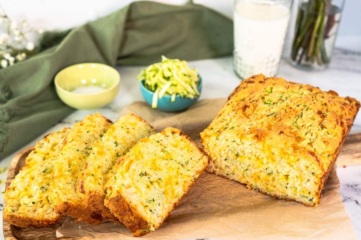 A loaf of sliced zucchini bread sits on parchment paper with a bowl of shredded zucchini and a glass of milk in the background—a delicious option for creative zucchini recipes.