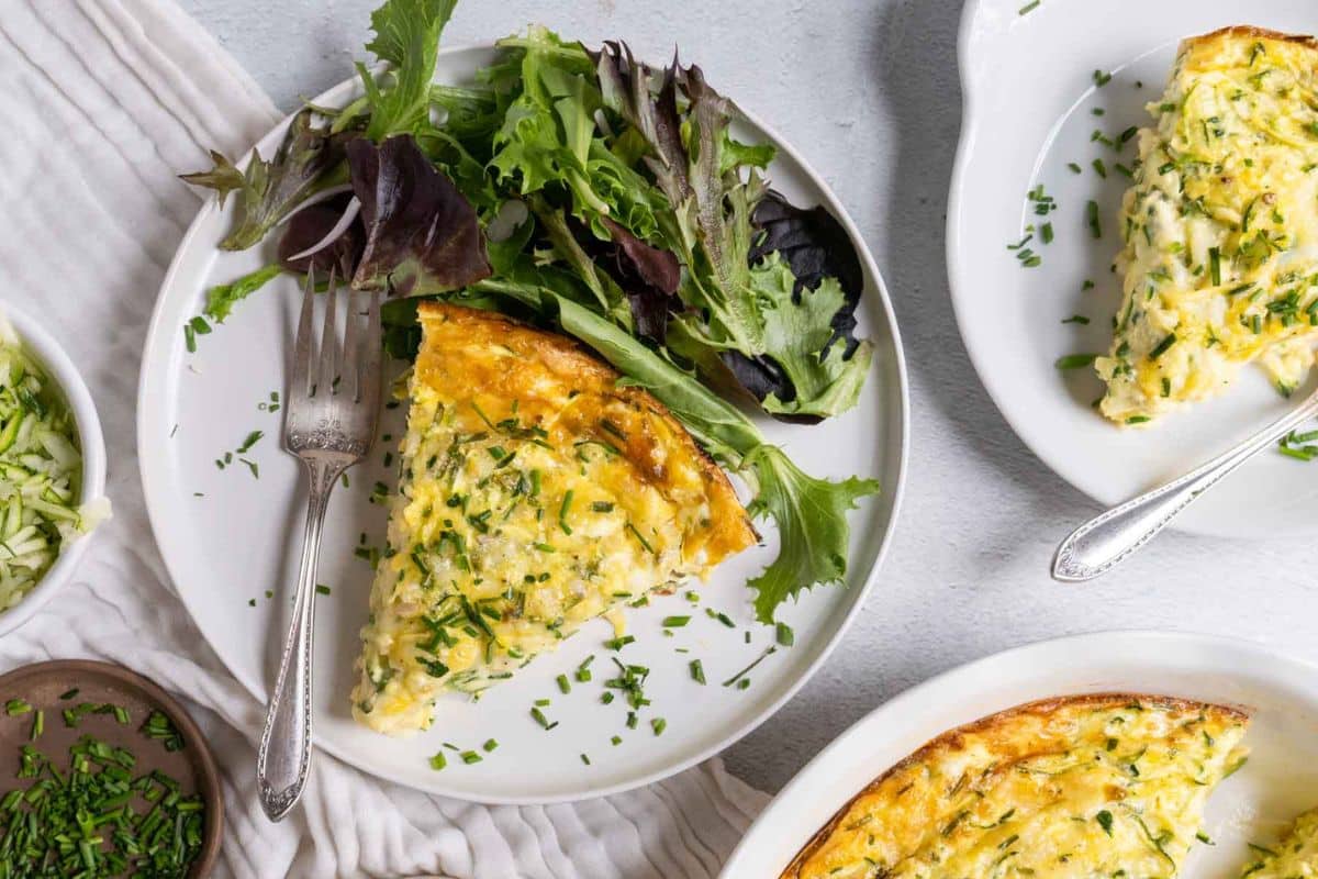 A slice of quiche, inspired by creative zucchini recipes and garnished with chopped chives, is served on a white plate alongside a small mixed green salad.