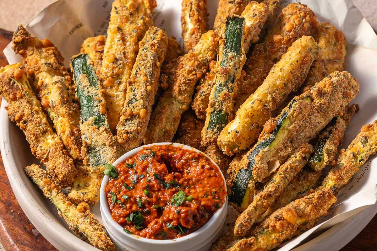 A bowl of breaded and baked zucchini fries is served with a small dish of chunky tomato dipping sauce—a delicious addition to your creative zucchini recipes collection.