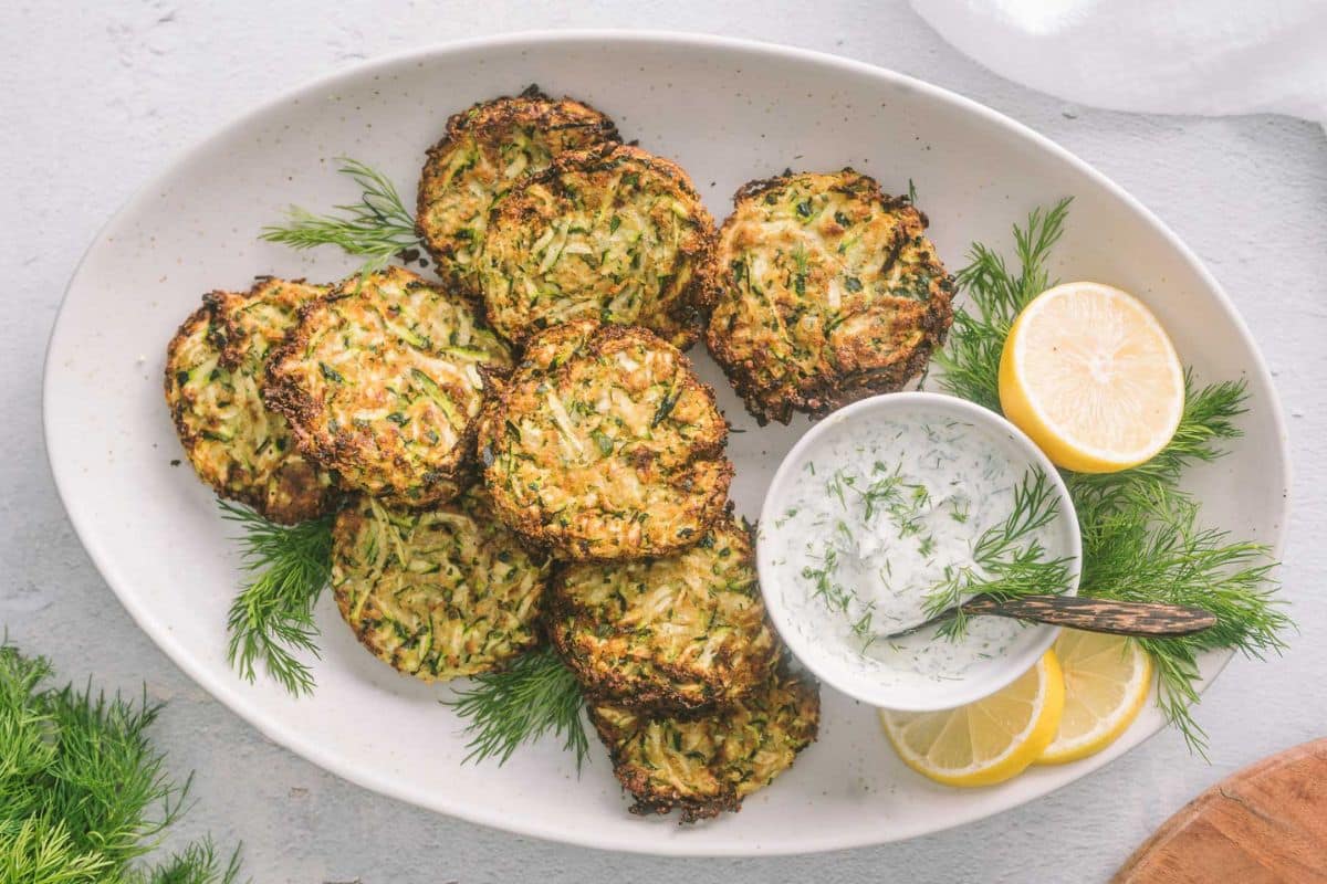 A plate of zucchini fritters garnished with fresh dill, served with a bowl of creamy dipping sauce and lemon slices—a delicious addition to your collection of creative zucchini recipes.