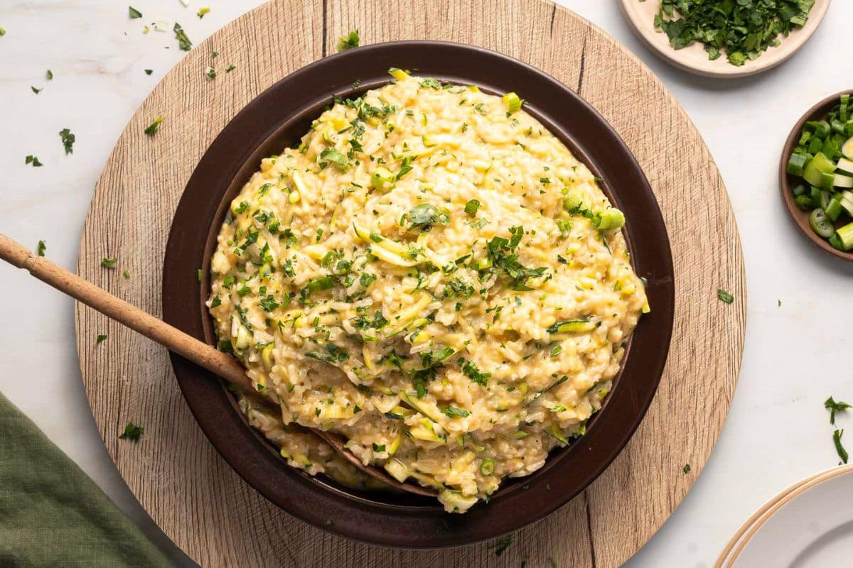 A bowl of creamy zucchini rice garnished with chopped herbs, with a wooden spoon resting inside, set on a wooden board—an inviting addition to your collection of creative recipes.