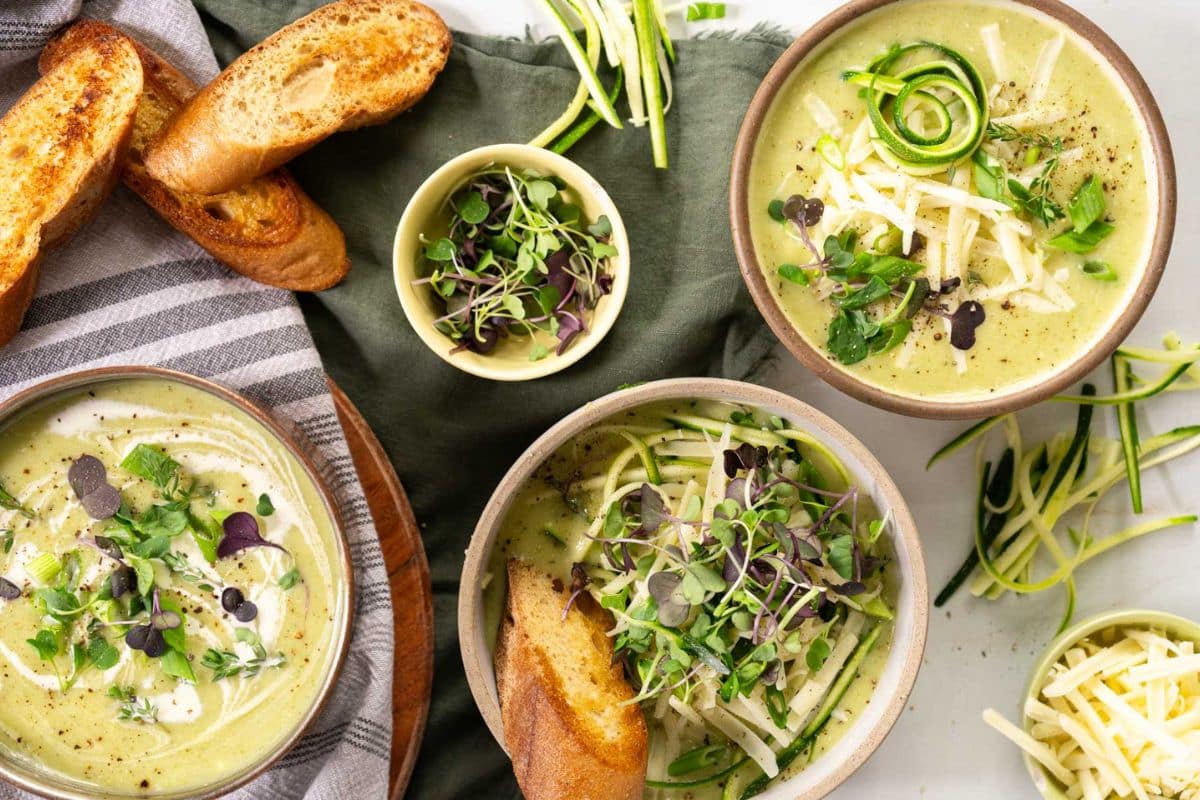 Three bowls of creamy green soup topped with shredded cheese, microgreens, and zucchini spirals—perfect for summer recipes or anyone seeking creative zucchini recipes—served with toasted baguette slices and bowls of garnishes.